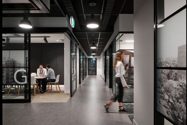 A modern commercial office interior viewed along a central corridor, with glazed meeting rooms on either side, a small meeting table with two people at left, black-framed glass partitions, dark exposed ceiling services, suspended circular lights, and polished grey flooring. Framed monochrome artwork is visible on the right wall.