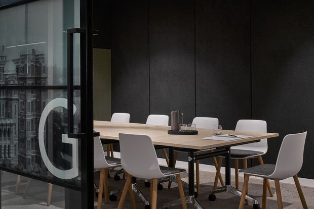 A contemporary meeting or conference room viewed through a glass partition, furnished with a long light-wood table and several white upholstered chairs. The room has dark charcoal wall panels, black-framed glazing with a large graphic letter, slim black table supports, and a restrained professional palette. An urban building is visible through the glazing.