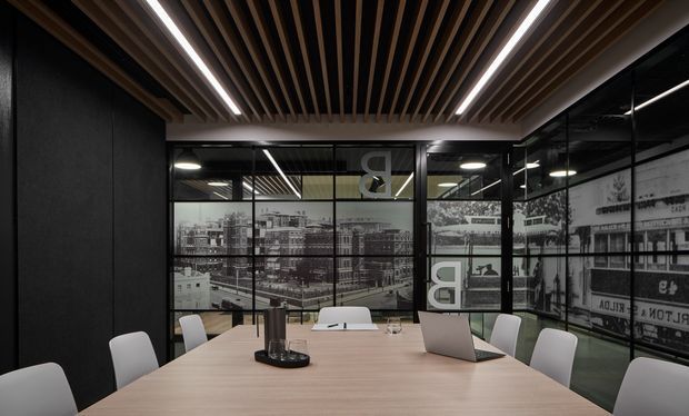 A contemporary office meeting room with a long light-wood conference table, white upholstered chairs, an open laptop, black-framed glass walls, and a dramatic black-and-white urban graphic film applied to the glazing. The room features a dark wall finish and a suspended slatted timber ceiling with integrated linear lighting, creating a refined, high-contrast commercial interior.
