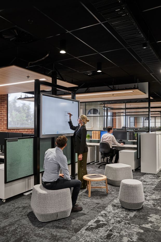 A contemporary open-plan commercial office interior with collaborative workstations, a presentation screen, glass partitions, exposed black ceiling services, timber acoustic ceiling panels, brick walls, and informal grey upholstered seating. Several people are using the workspace, with desks and monitors arranged around a central collaboration area.