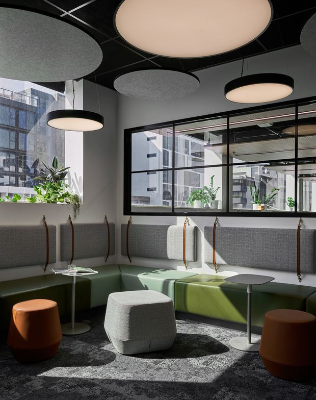 Contemporary commercial lounge or breakout area with a curved, upholstered green banquette wrapping around the room, small pedestal tables, sculptural stools, and a large central ottoman. The space has a dark ceiling with oversized circular acoustic pendant fixtures, grey acoustic wall panels, black-framed internal glazing, abundant plants, and strong natural light. The setting appears to be an office or hospitality-style communal interior.