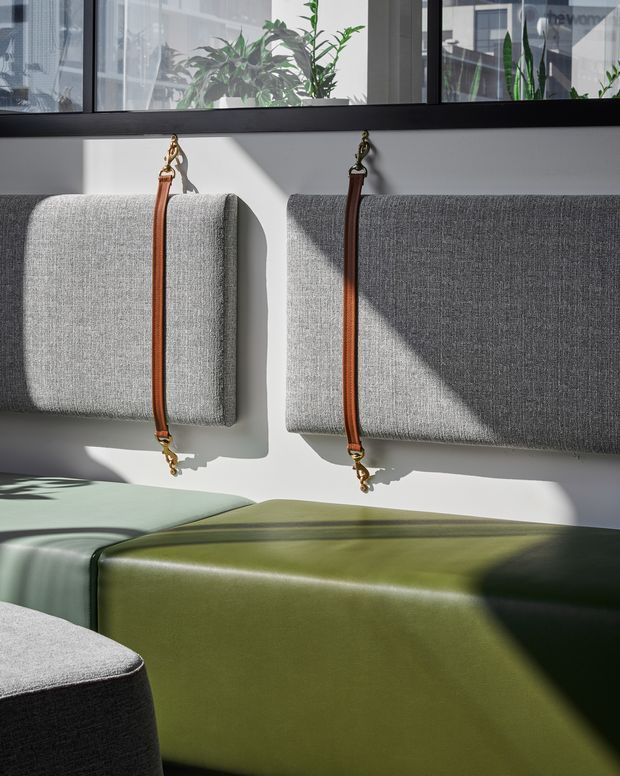 A contemporary commercial-style interior with modular upholstered seating in grey, sage green, and olive tones. Two grey wall-mounted upholstered panels are secured with vertical brown leather straps and brass-toned hardware. Strong sunlight creates geometric shadows across the seating and white wall, while indoor plants are visible behind a glazed upper partition.