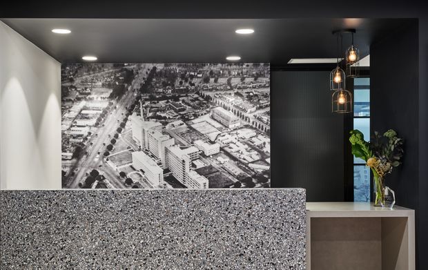A dark, refined kitchen or bar area featuring a large speckled stone island or counter in the foreground, dark cabinetry and ceiling, recessed downlights, and a cluster of hanging glass pendant lights. A dramatic black-and-white aerial city photograph forms a feature wall, with a small floral arrangement beside a window or glazed opening.