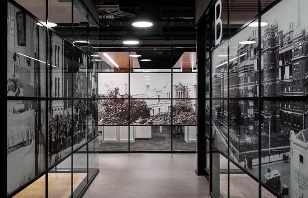 A modern commercial interior viewed along a glazed corridor or office entry zone. Black-framed floor-to-ceiling glass partitions are covered with large monochrome architectural and city imagery, opening onto a contemporary workspace with desks and office chairs. The exposed black ceiling contains recessed services and circular pendant-style lights, while the floor is finished in a muted grey-brown surface. The space has a sophisticated, graphic, high-contrast office fit-out.