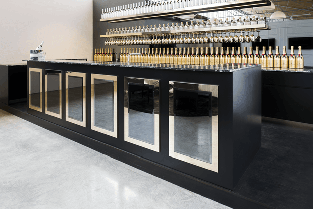 A sophisticated commercial bar or tasting-room interior dominated by a long matte black counter with multiple gold-trimmed mirrored inset panels. The counter has a dark stone-look top and is backed by extensive illuminated shelving displaying rows of bottles and glassware. Polished concrete flooring, dark cabinetry, and monochromatic finishes create a refined, contemporary atmosphere.