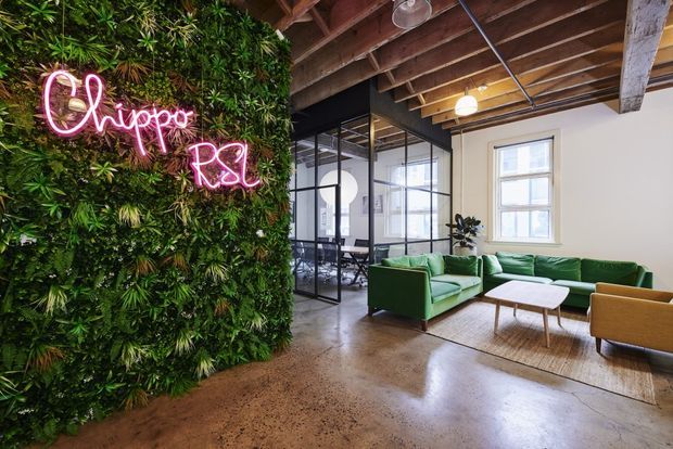 A stylish commercial office lounge with a lush preserved-looking green wall and pink neon signage reading “Chippy RSL.” The space features polished concrete flooring, exposed timber roof beams and services, black-framed glass meeting-room partitions, a green sectional sofa, mustard lounge chairs, a white coffee table, and a neutral area rug. Large windows provide abundant natural light.