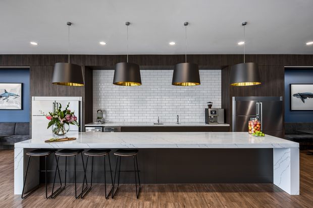 A spacious contemporary kitchen centred on a large white marble-look island with seating for four. The room features dark wood cabinetry, a white subway-tile splashback, stainless-steel appliances, timber flooring, and four oversized pendant lights with metallic gold interiors. Blue accent walls and framed artwork are visible at either side.