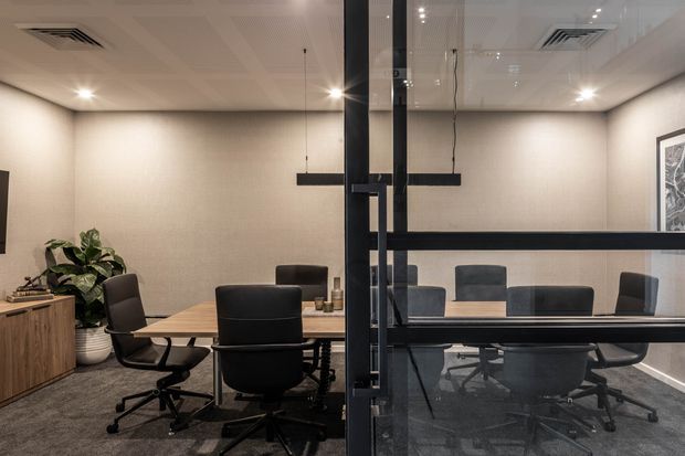 A contemporary meeting room viewed through a glazed partition, with a long timber conference table surrounded by black office chairs. The room has grey carpet, warm neutral textured walls, a suspended acoustic-panel ceiling with recessed lighting, black-framed glazing and doors, built-in timber storage, indoor planting, and framed artwork.