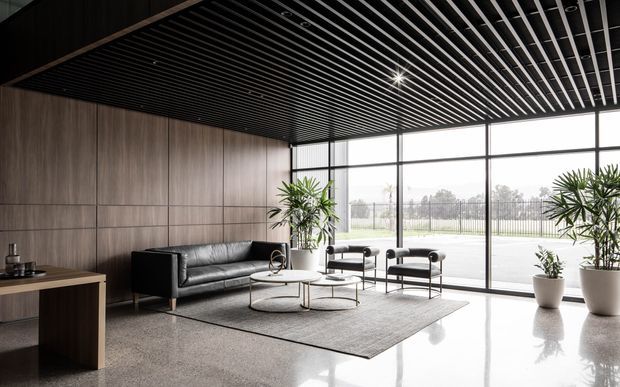A sophisticated contemporary lobby or reception lounge with dark timber wall panelling, a black slatted ceiling, full-height glazing, polished light flooring, a black leather sofa, sculptural lounge chairs, round nesting coffee tables, and indoor potted plants. The space is bright despite its dark ceiling and feature wall, with a refined, minimalist commercial interior aesthetic.