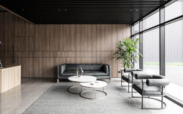 A refined contemporary reception or lounge area with dark timber wall panelling, polished concrete or terrazzo flooring, a black slatted ceiling, expansive black-framed glazing, and coordinated black leather seating around nested round coffee tables. A large indoor plant adds a natural accent, while the restrained palette and strong linear detailing create a sophisticated commercial interior.