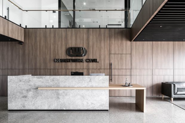 A refined commercial reception area with a large stone-fronted reception desk, warm vertical timber wall panelling, integrated branding, glass balustrades overlooking an upper level, and a dramatic black slatted ceiling feature. A small seating area is visible to the right, and the floor appears to be polished terrazzo or concrete.
