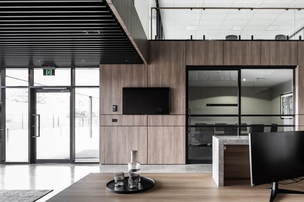 A contemporary commercial interior, likely an office reception or lounge, featuring a wood-panelled built-in media wall, black-framed glass meeting room, reception counter, black ceiling slatted feature, polished concrete floor, and a large desk or table in the foreground. The space has a restrained neutral palette with strong black and timber contrasts.