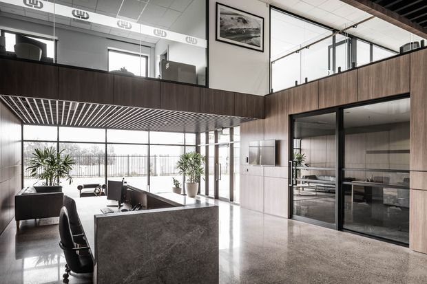 A bright, contemporary commercial office reception and open-plan workspace with a polished concrete floor, dark stone reception desk, extensive black-framed glazing, timber-look wall panelling, and a mezzanine level with exposed ceiling slats. Large indoor plants and glass-partitioned meeting rooms contribute to an airy, professional atmosphere.