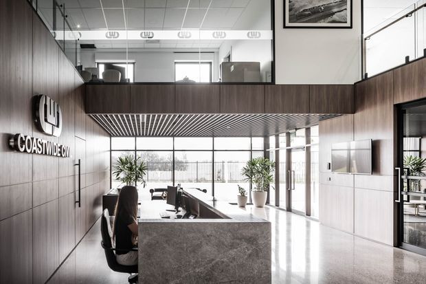 A sophisticated double-height commercial reception and workspace with dark timber wall panelling, a stone reception counter, expansive glazing, indoor plants, and a mezzanine-level office overlooking the lobby. The space has a polished, contemporary corporate aesthetic with strong natural light and linear ceiling detailing.