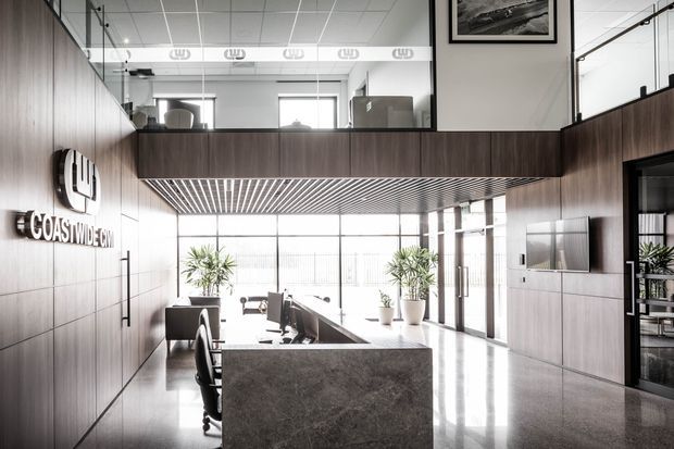 A bright contemporary commercial reception and office interior with a double-height void. Dark timber wall panelling and built-in joinery frame a stone reception counter, while extensive glazing, polished flooring, slatted ceiling detailing, indoor plants, and an upper-level workspace create an open professional environment. The Coastwide sign identifies the reception area.