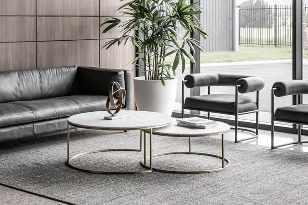 A refined contemporary living room with a black leather sofa, matching upholstered lounge chairs, nested round coffee tables with slim metallic frames, and a large neutral rug. Timber-toned wall panelling, floor-to-ceiling glazing, and a large indoor plant create a warm, light-filled setting.