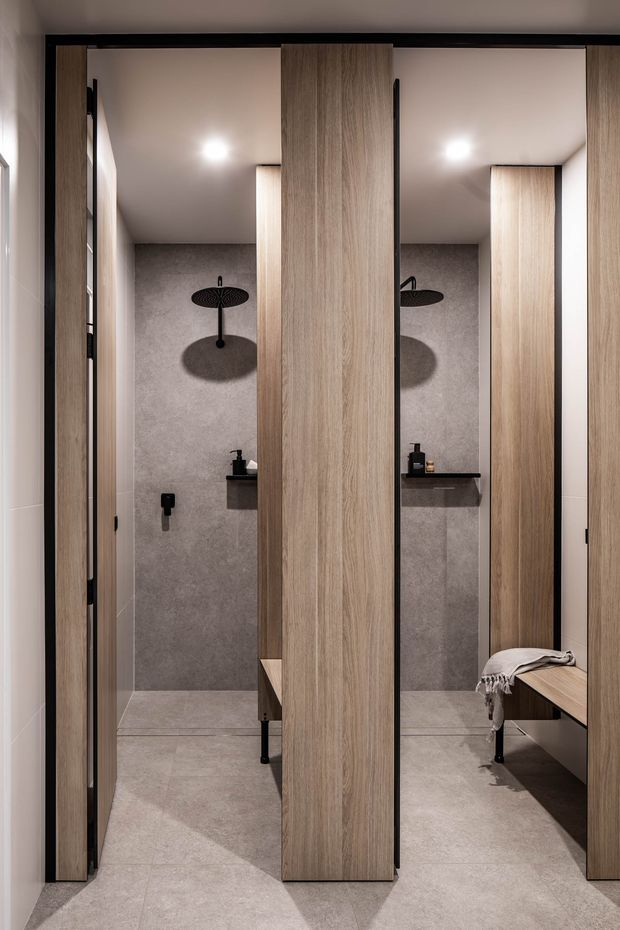 A contemporary bathroom with two adjacent open shower cubicles, separated by full-height light timber privacy screens. Each shower has grey stone-look wall and floor tiles, black round rain shower fittings, recessed lighting, and a slim black wall shelf. A timber bench with a folded throw is visible on the right, and the overall design is warm, minimalist, and highly refined.