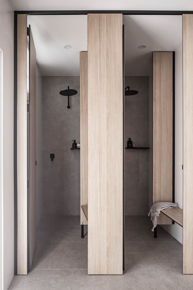 A compact contemporary bathroom with two open shower cubicles framed by pale timber-look vertical partitions. Each shower has dark grey wall and floor tiles, matte black shower fittings and recessed shelving, while integrated timber benches and cabinetry add warmth. The symmetrical, space-efficient layout has a clean minimalist appearance.