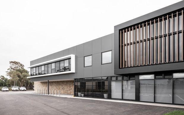 Contemporary multi-storey commercial building exterior with a predominantly charcoal and grey facade, extensive glazing, and a prominent upper-level screen of vertical timber-toned fins. A lighter projecting box frames the upper-floor windows on the left, while textured stone cladding defines the lower section. The building fronts a paved parking area and is photographed from outside.