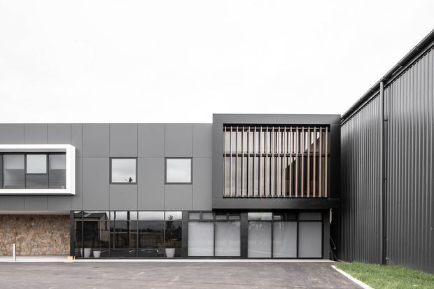 Contemporary multi-level building facade with dark grey vertical metal cladding, charcoal panel cladding, and a projecting upper-level volume screened by closely spaced timber-toned vertical battens. Large glazed openings, a white-framed projecting window, paved forecourt, and a narrow strip of lawn are visible under an overcast sky.