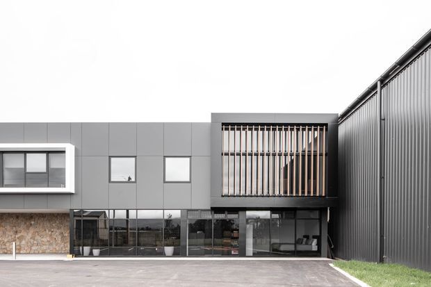 Contemporary multi-level building facade with dark grey vertical metal cladding, smooth grey panel cladding, large glazed openings, and a projecting upper-level balcony or screened enclosure. The upper opening features closely spaced timber-toned vertical slats, while the lower level has extensive glazing and a stone-clad wall section. A paved forecourt and narrow strip of lawn are visible.