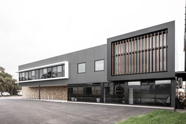 Contemporary two-storey commercial building with a dark grey panelled facade, extensive ground-floor glazing, and a prominent upper-level screened window bay featuring vertical timber-toned fins. A lighter grey upper volume projects over a stone-textured lower wall, creating a crisp modern office or commercial frontage. The building is viewed externally from a paved forecourt, with limited landscaping at the edge.