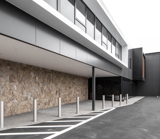 Exterior architectural view of a contemporary multi-level building with a projecting upper volume, extensive dark-framed glazing, charcoal cladding, and a textured natural stone wall at the sheltered entry. A covered pedestrian zone with bollards and diagonal road markings fronts the building.