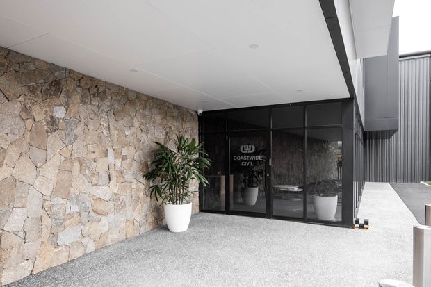 A contemporary commercial building entrance beneath a broad white soffit. The facade combines irregular natural stone walling with black-framed glazed entry doors and dark vertical metal cladding. Potted plants flank the entrance, and a textured light gravel or aggregate forecourt leads toward the doorway. The glass bears Coastwide Civil signage.