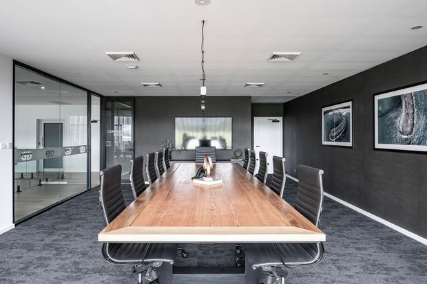 Contemporary corporate conference room with a long timber meeting table, black leather office chairs, dark textured walls, carpeted flooring, framed artwork, glass-partitioned offices, and a wall-mounted display. Suspended linear lighting and recessed ceiling services reinforce the commercial workplace setting.