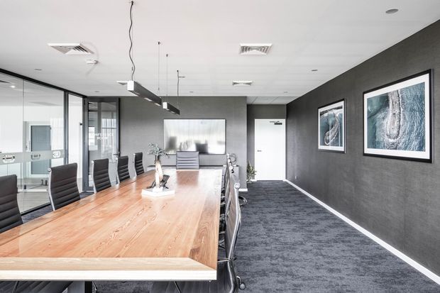 Contemporary commercial boardroom with a long solid timber conference table, black executive chairs, dark textured walls, carpeted flooring, framed artwork, glass partitions, and linear pendant lighting. The room has a clean, professional and moderately high-contrast fit-out.