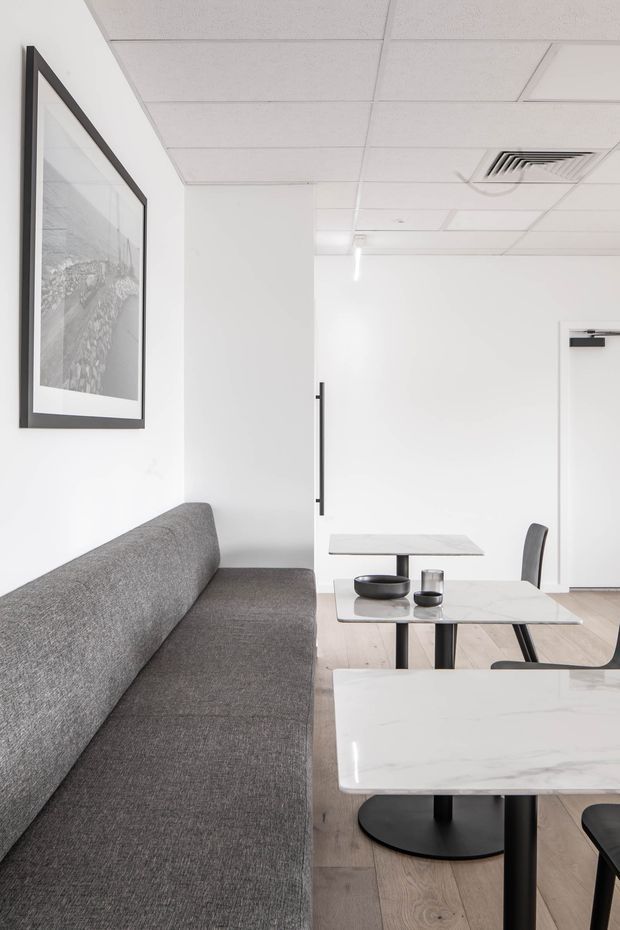 A bright, minimalist commercial-style interior with a long grey upholstered bench sofa along a white wall, two small marble-look tables with black pedestal bases, and a compact dining or meeting table with black chairs. Pale timber flooring, white walls, a suspended acoustic tile ceiling, recessed air-conditioning vent, and framed monochrome artwork create a clean, refined setting.