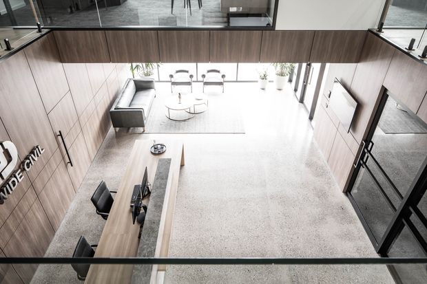 High-angle view of a contemporary open-plan commercial interior, likely a reception or showroom space. The room features extensive warm timber wall panelling and cabinetry, a long reception/work counter with office chairs, a grey lounge sofa and coffee tables, light carpet, full-height glazing, and a bright, double-height volume with an upper-level glazed edge.