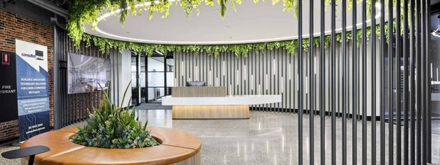 A contemporary commercial reception lobby with a polished light floor, curved timber reception desk and matching circular planter seating, lush greenery, a large suspended oval ceiling feature, vertical metal screening, exposed brick, and abundant glazing. The space appears refined and biophilic, with a visible reception counter and branding signage.
