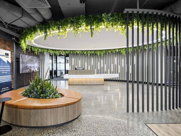 Contemporary commercial reception lobby with a curved timber-clad planter bench, polished terrazzo flooring, a minimalist reception desk, vertical dark screening battens, exposed services, and abundant suspended greenery surrounding a circular ceiling feature.