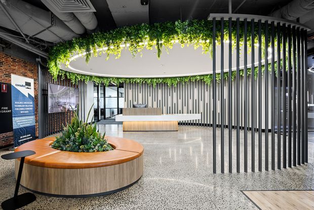 A contemporary commercial reception and waiting area with a large curved timber-clad built-in bench surrounding a planted central bed, a matching reception desk, polished terrazzo flooring, vertical metal screening, exposed services, brickwork, and a dramatic circular ceiling feature with hanging greenery.
