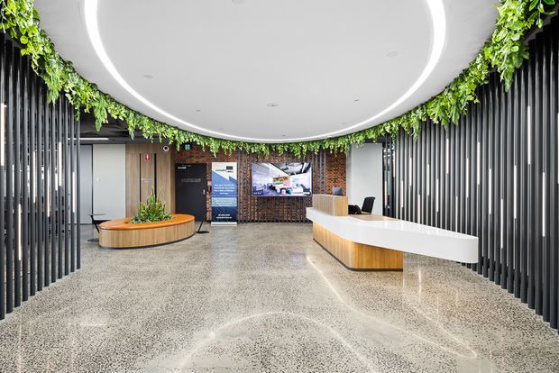 A contemporary commercial reception lobby with a curved white ceiling featuring integrated circular lighting, dark vertical slatted wall screens, hanging greenery, terrazzo flooring, timber accents, and a long white reception desk. A rounded timber planter bench and reception signage create a polished, welcoming entry environment.
