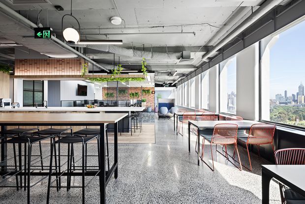 Bright industrial-style commercial interior with a large communal island and bar stools in the foreground, a kitchen or service counter beyond, and café-style tables and chairs arranged beside expansive windows. The space has an exposed concrete ceiling with visible services, polished terrazzo-like flooring, black metal framing, and city views through the glazing.