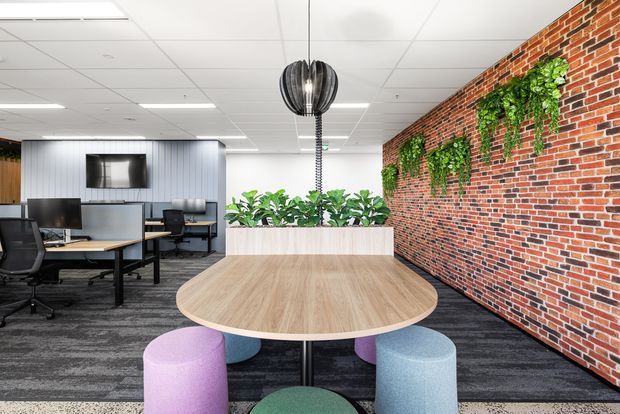 A contemporary open-plan office with a large rounded timber meeting table surrounded by colourful upholstered stools. The space includes workstations with computers, a dark carpeted floor, a suspended sculptural pendant light, a brick feature wall with hanging greenery, and a raised planter dividing the meeting area from the work zone.