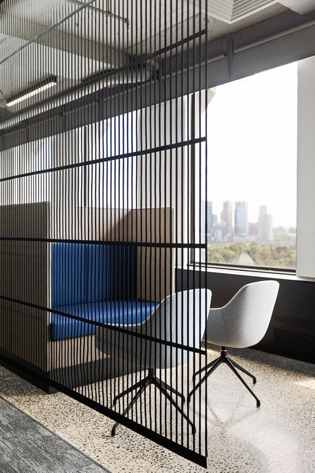 A contemporary office or commercial interior with a black metal vertical-rod partition creating a semi-private seating nook. The space includes a vivid blue upholstered bench, two light grey molded lounge chairs, exposed dark structural elements and services, large windows with an urban skyline view, and a polished terrazzo floor.