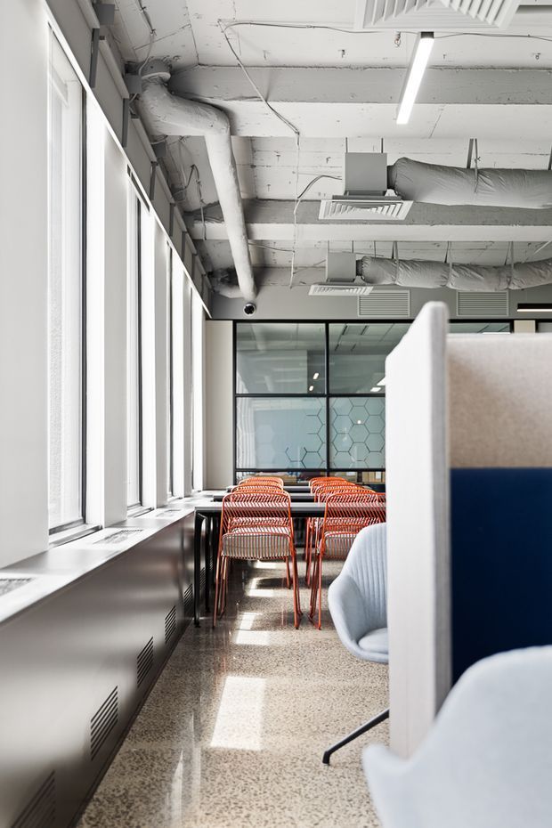 Bright contemporary commercial workspace with large windows, exposed white-painted services and ductwork, linear LED lighting, terrazzo flooring, orange wire-frame dining or breakout chairs, and blue-and-white upholstered partitioned seating. Glazed meeting-room partitions with geometric privacy film are visible at the rear.