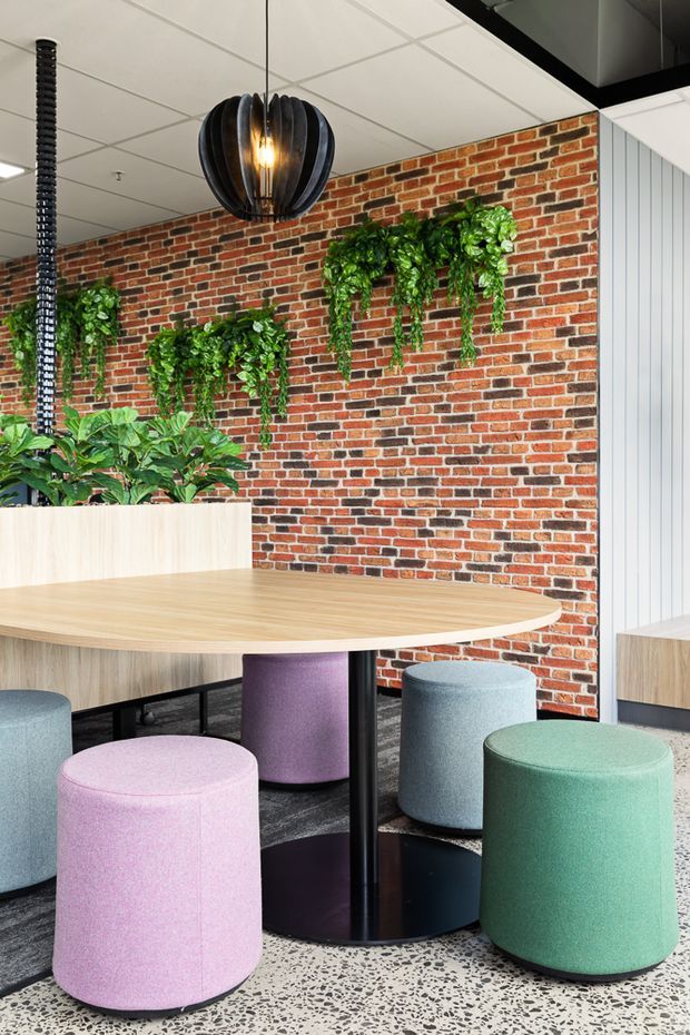 A vibrant contemporary communal dining or breakout area with a large rounded timber table, colourful upholstered cylindrical stools, a red exposed-brick feature wall with cascading greenery, terrazzo flooring, and sculptural pendant lighting. The space has a playful, biophilic commercial-interior character.