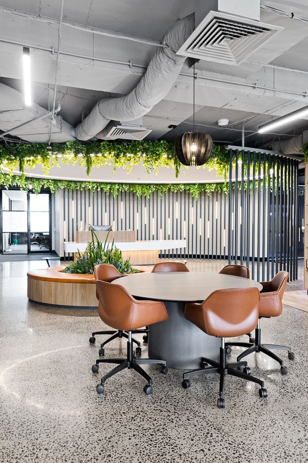 A contemporary commercial office breakout or meeting area with a large oval table surrounded by tan leather swivel chairs. The space features polished terrazzo flooring, curved timber-look built-in seating, vertical dark screening slats, abundant suspended greenery, exposed painted services and ductwork, and decorative pendant lighting.