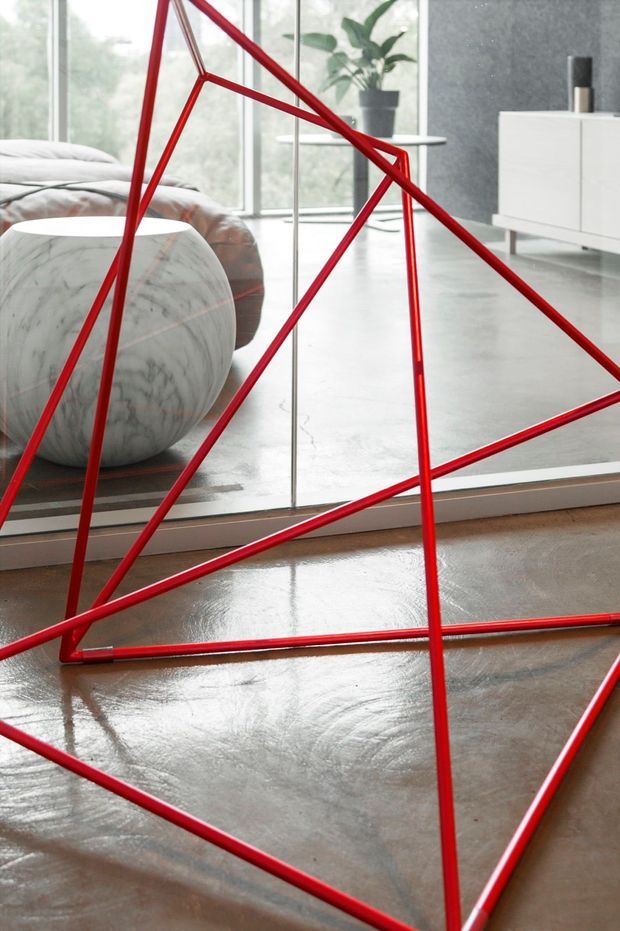 A contemporary living room viewed through a large mirror, featuring a dramatic red geometric metal sculpture or room-divider structure in the foreground. The space includes polished concrete flooring, floor-to-ceiling glazing, a white marble-like rounded lounge chair, a grey sectional sofa, a white minimalist sideboard, and a potted plant. The image has a clean, gallery-like aesthetic with strong red linear accents.
