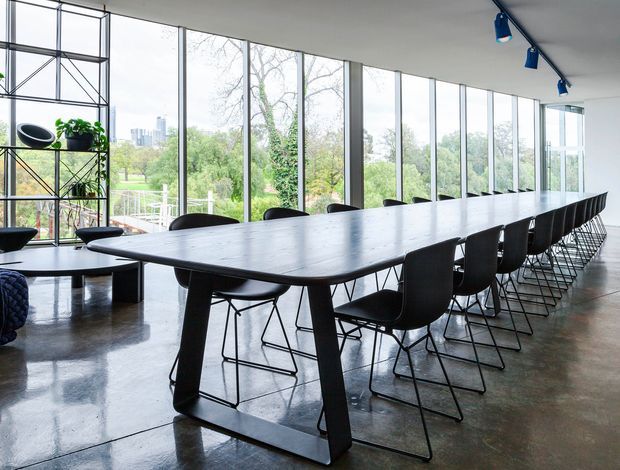 Bright contemporary meeting or dining space with a long dark timber table and numerous black wire-frame chairs. Full-height glazing overlooks landscaped greenery and a distant urban skyline, while polished concrete floors, a simple white ceiling, and exposed black shelving with plants create a refined industrial feel. Blue directional track lighting adds a small accent.