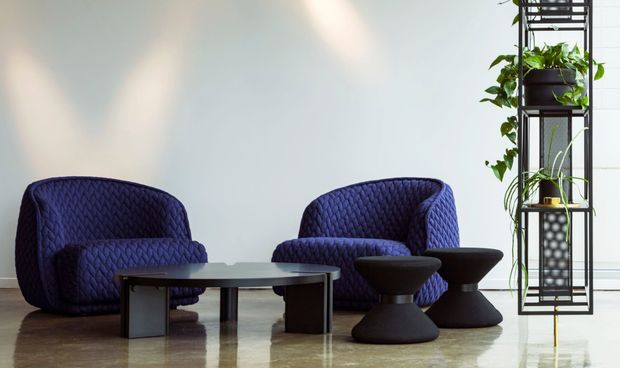 A refined lounge setting with two deep navy quilted lounge chairs, a low rectangular coffee table, and sculptural black side tables. A tall black metal shelving unit with cascading green plants stands to the right. The room has a pale wall, polished reflective flooring, and a calm, contemporary arrangement.