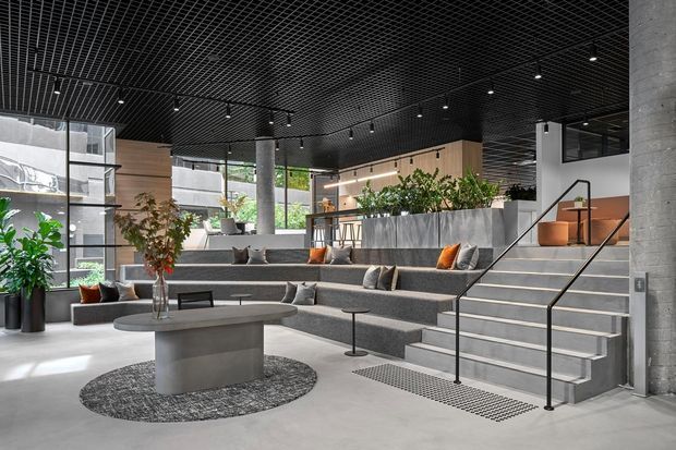A spacious contemporary commercial-style lounge and reception area with tiered upholstered seating, a low round coffee table, large planters, exposed concrete columns, timber wall panels, and broad stair steps with black handrails. The ceiling features a black exposed grid with track lighting, while extensive glazing brings in daylight and greenery.