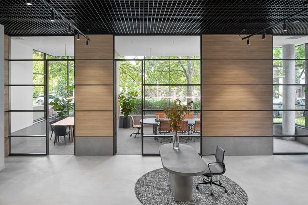 Contemporary office interior with a central meeting table and office chair in the foreground, surrounded by glazed meeting rooms and workspaces. The space features full-height black-framed glazing, warm timber wall panelling, a polished concrete floor, a dark exposed grid ceiling with track lighting, and abundant greenery visible through the windows.