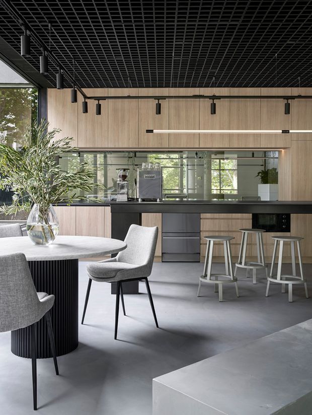 Contemporary open-plan kitchen and dining area with full-height timber-toned cabinetry, a long black island bench, integrated appliances, and a mirrored splashback. A round dining table with upholstered chairs occupies the foreground, while stools line the island. The space features a black exposed grid ceiling with suspended cylindrical pendant lights, large windows, and a polished concrete floor, creating a refined industrial-modern atmosphere.