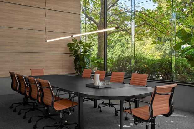 A modern conference room with a large dark meeting table surrounded by orange-brown upholstered office chairs. A slim linear pendant light hangs above the table, while a warm wood-panelled wall and expansive floor-to-ceiling black-framed glazing overlook dense greenery. Potted plants add an organic element to the refined commercial interior.