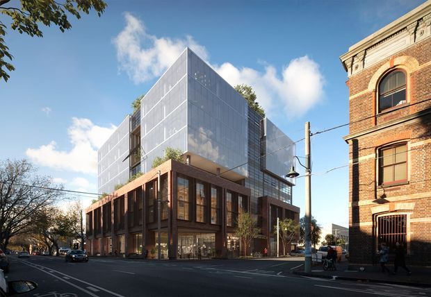 Street-level exterior view of a contemporary multi-storey urban building, combining a transparent glass-and-metal upper volume with a lower podium of dark brick, timber-toned framing and extensive glazing. It sits beside a historic red-brick masonry building on a tree-lined city street under a bright blue sky.